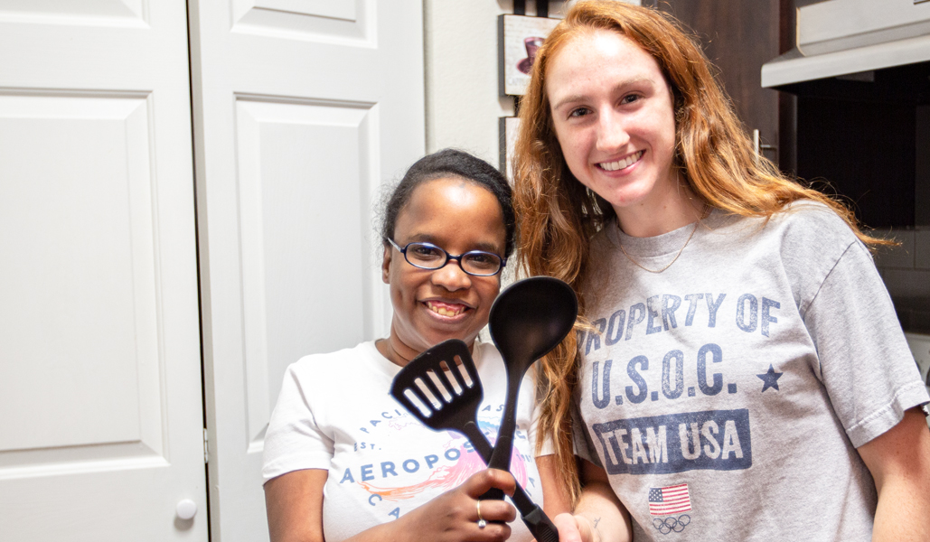 Two people smiling and holding cooking utensils in a kitchen.